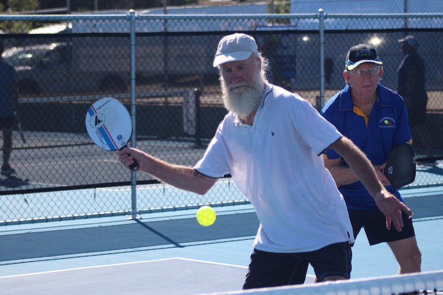 Two older men playing pickleball with one man attempting to strike the plastic ball with his paddle