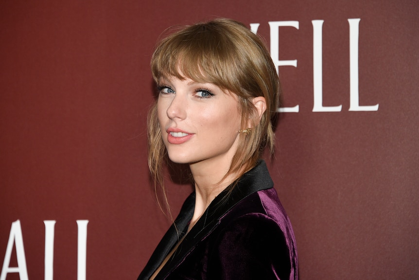 Taylor Swift smiles at the camera in front of a maroon backdrop. 