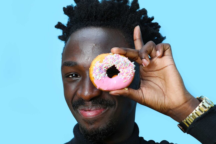 A man smiles as he holds up a pink iced doughnut to his eye, one of many doughnut varieties.