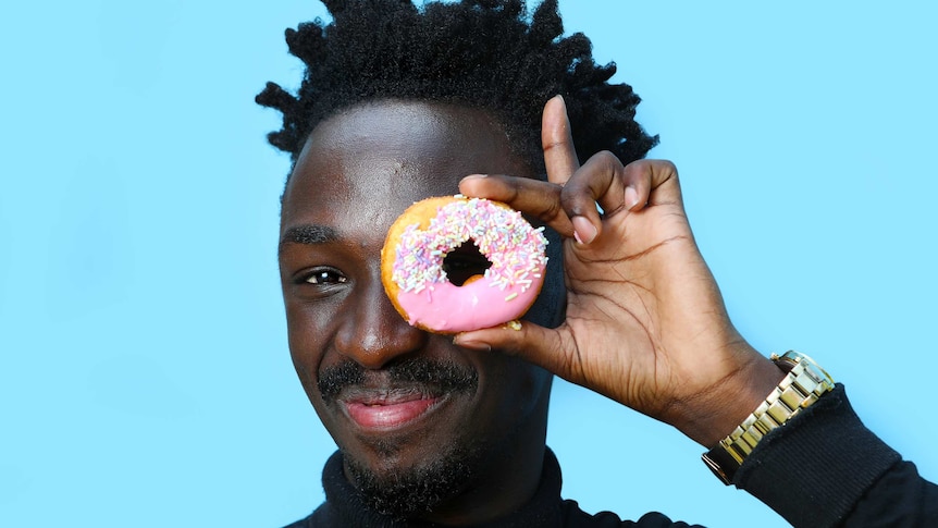 A man smiles as he holds up a pink iced doughnut to his eye, one of many doughnut varieties.