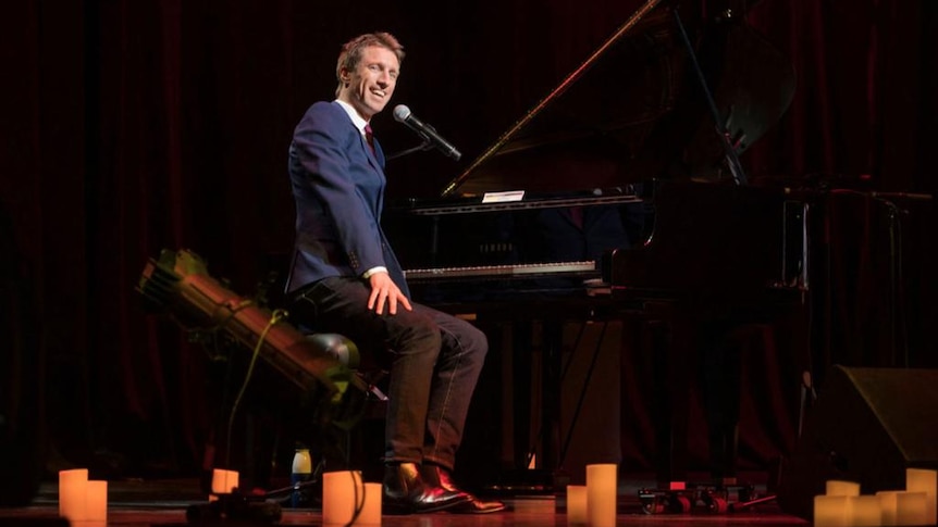 Sammy J on stage sitting at a piano, candles at his feet, wearing a blue suit