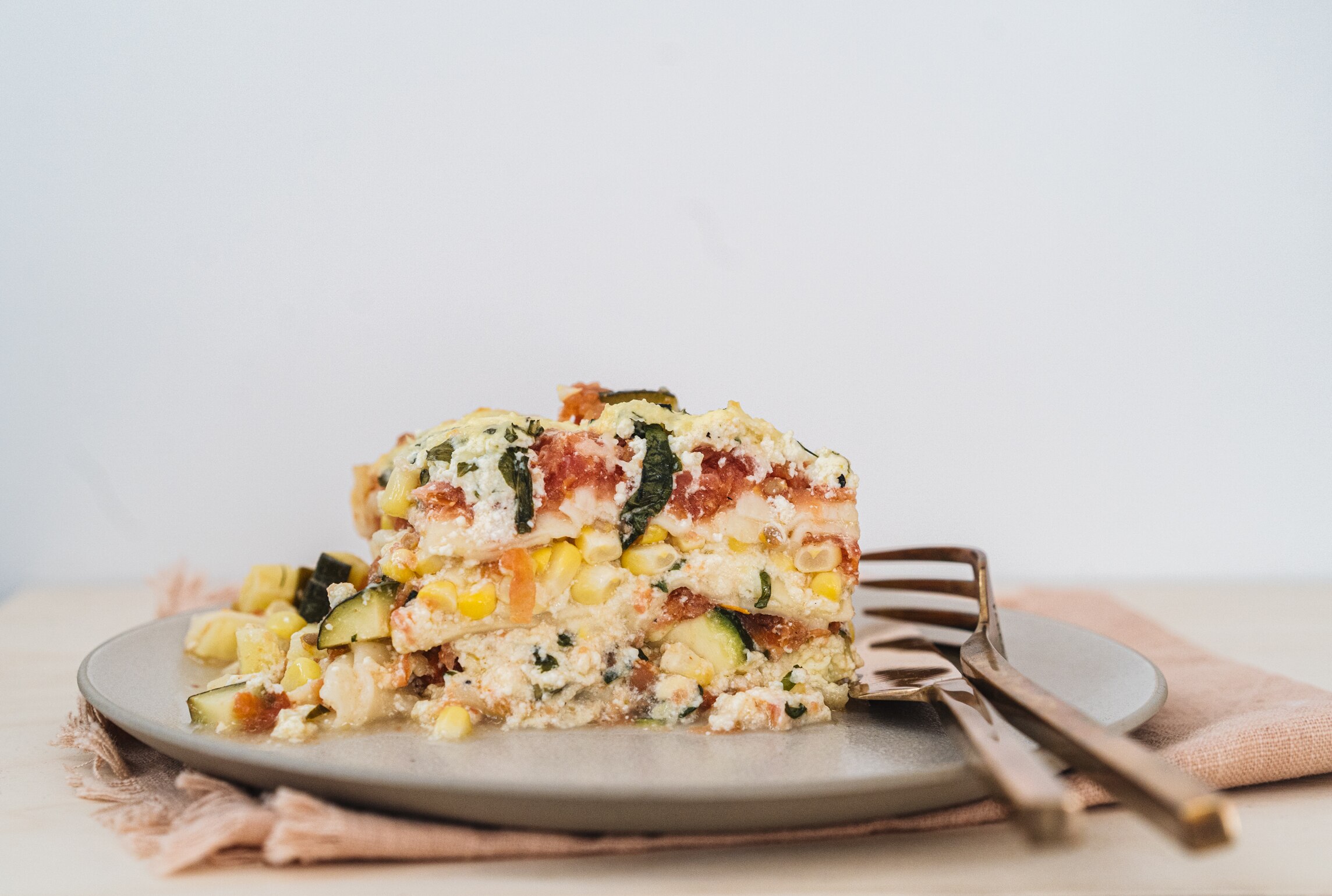 A slice of summer lasagne on a grey plate with two forks. It has layers of fresh corn, grated tomato, zucchini and ricotta.