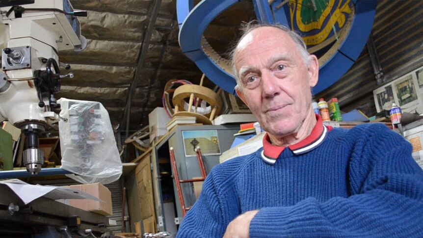 An older man with thin grey hair sits in a workshop, surrounded by machinery and cut outs of round sundials stacked to the roof.