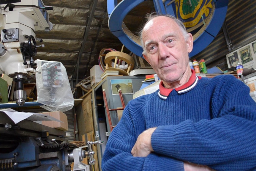 An older man with thin grey hair sits in a workshop, surrounded by machinery and cut outs of round sundials stacked to the roof.