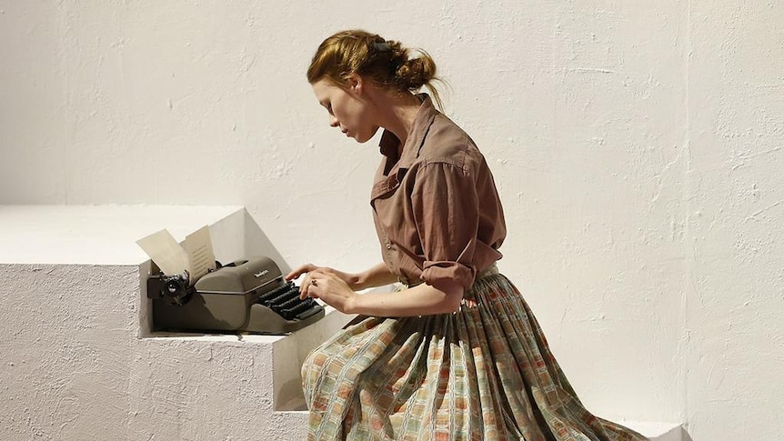 A woman in a brown shirt and floral skirt sits on some stairs with a typewriter