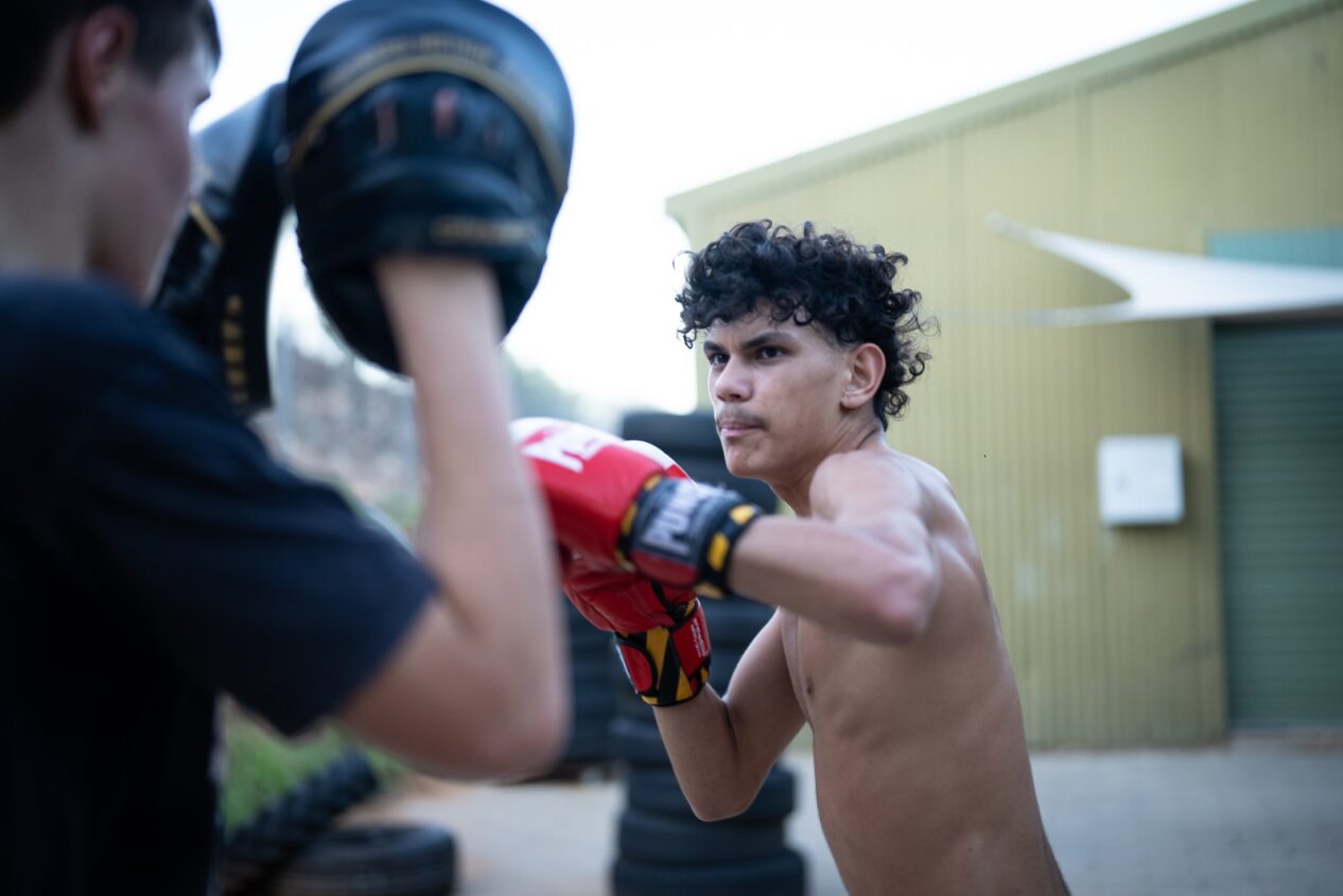 Teenager with shirt off prepares to throw a punch at pads held up in front of him by another teenager