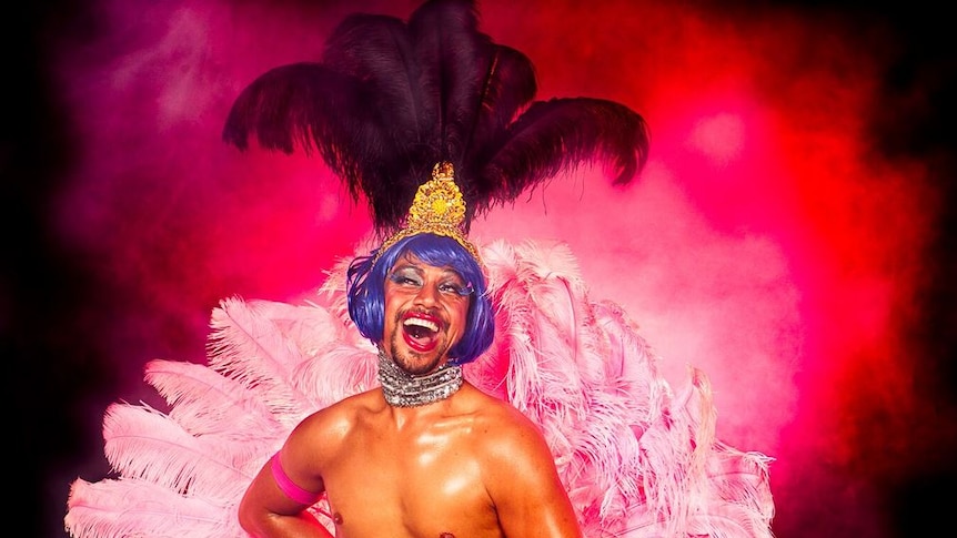 A shirtless man with a corset, purple wig and a huge pink feather fan.