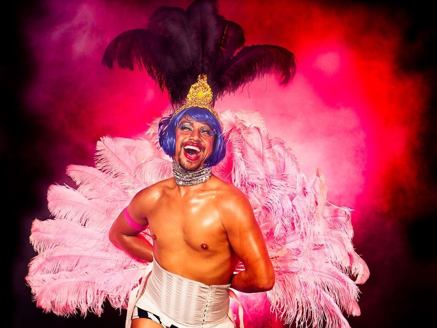 A shirtless man with a corset, purple wig and a huge pink feather fan.