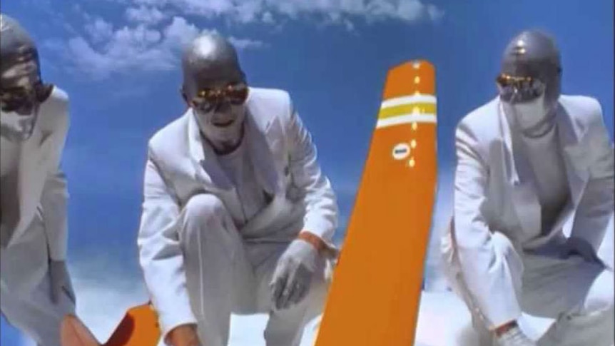 Three men in white suits with silver face paint hold a large orange model aeroplane