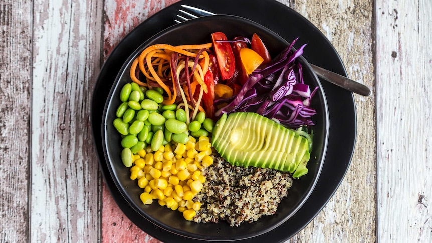 A black bowl full of quinoa, avocado, edamame, tomatoes, corn, red cabbage and carrots sits on a wooden table.