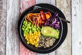 A black bowl full of quinoa, avocado, edamame, tomatoes, corn, red cabbage and carrots sits on a wooden table.