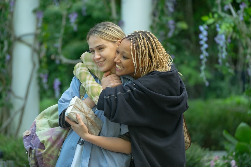 Black woman in her 20s with bleached braids embracing white women in her 20s wearing a blue top. 