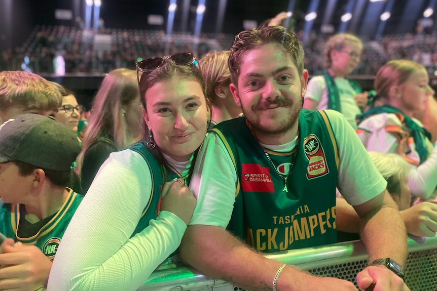 A young woman and man in a JackJumpers basketball jersey smile. They are at a barrier with a full stadium behind them.