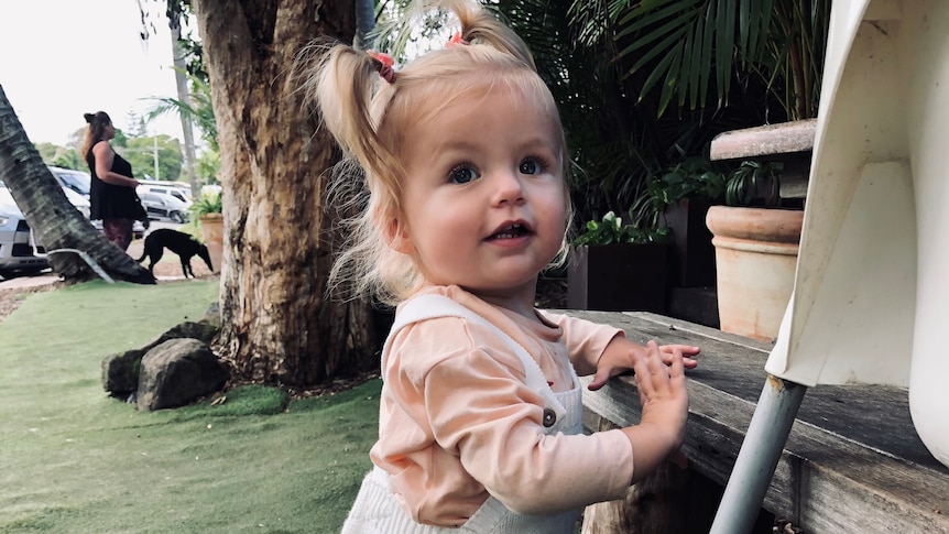A toddler with blonde hair in pigtails standing outside.
