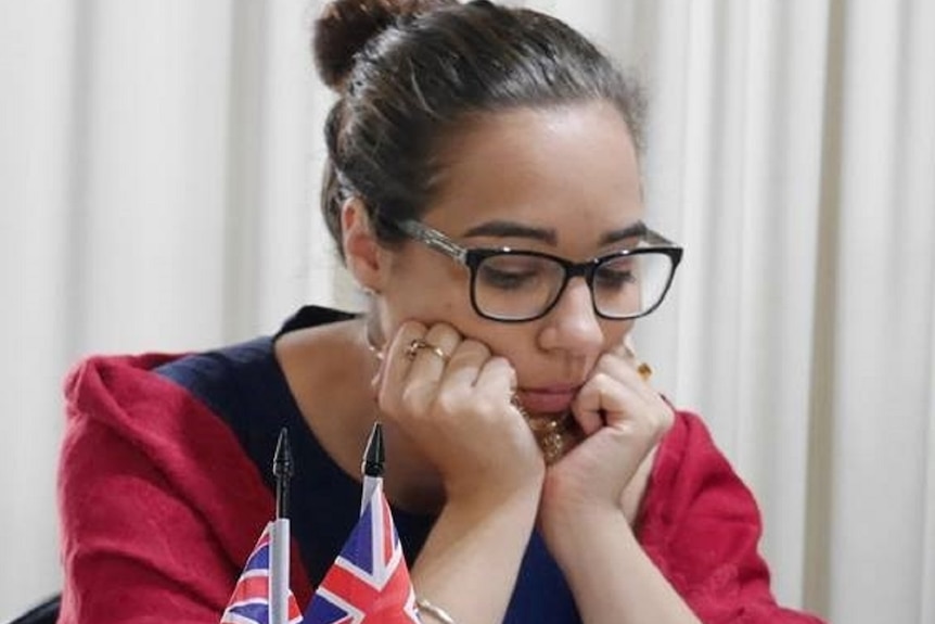 A woman in a red jacket sitting at a table in front of a chess board with her hands on her chin