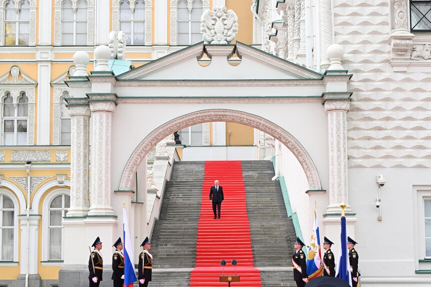 A small older white man in a suit walks down a red carpet on a long flight of stone, grey stairs.