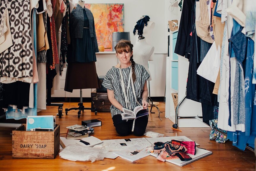 Designer Kelli Donovan kneeling in her studio surrounded by colourful items of clothing.