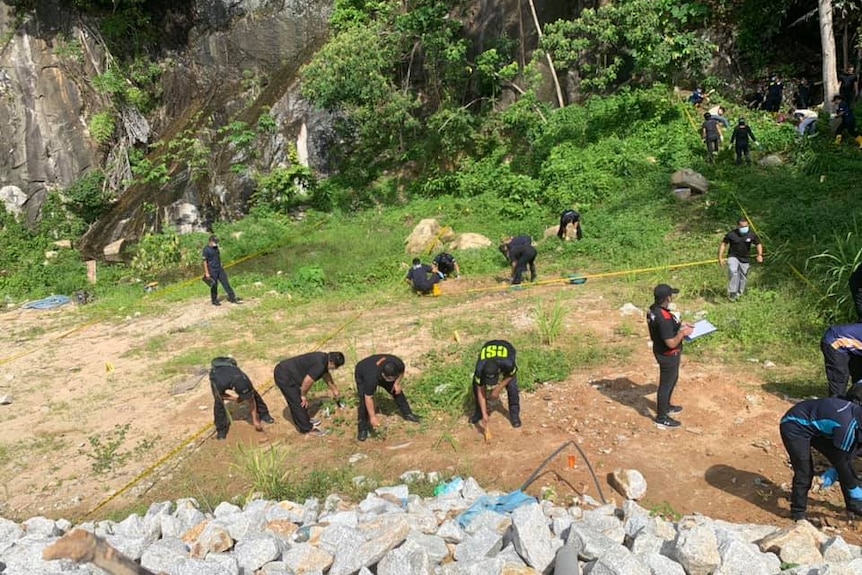 Police search a grassed area next rocks and trees