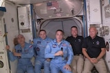 Five men in a spaceship smile at the camera
