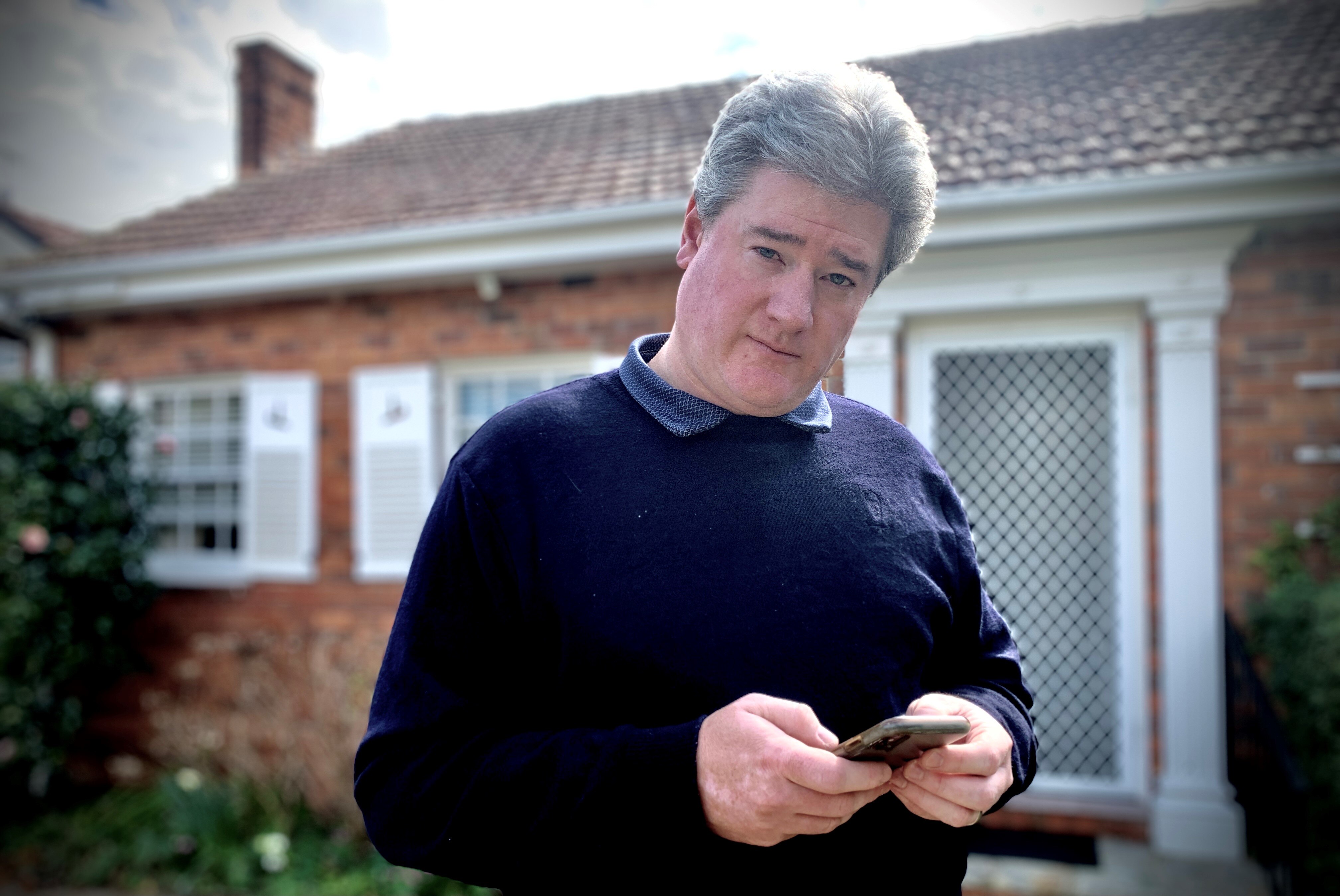 Simon Cowan stands in front of house holding his mobile phone.