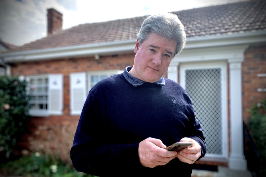 Simon Cowan stands in front of house holding his mobile phone.