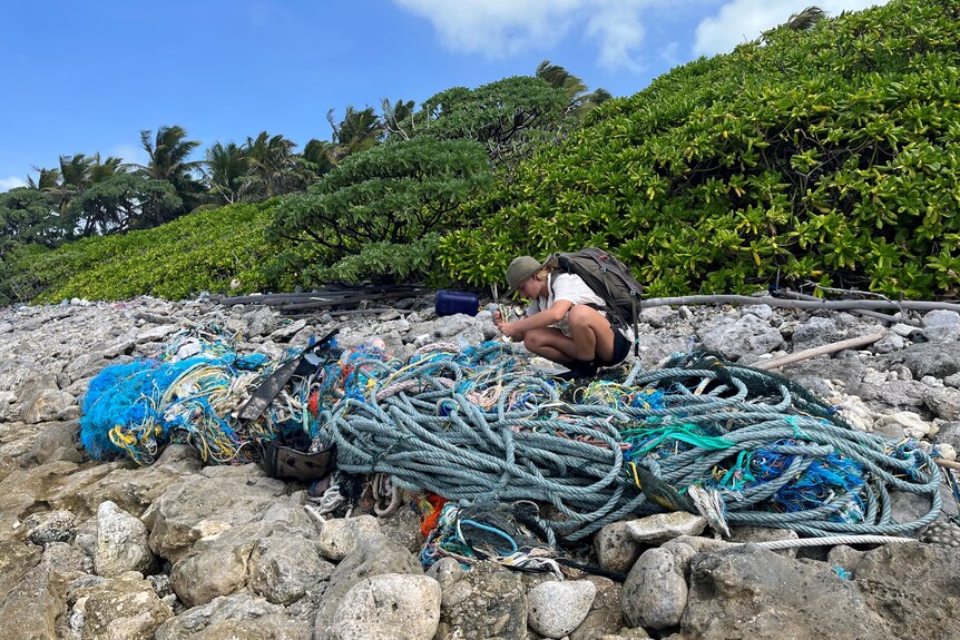 A woman kneels on boulders near a rugged incline, sorting through a large pile of rope.