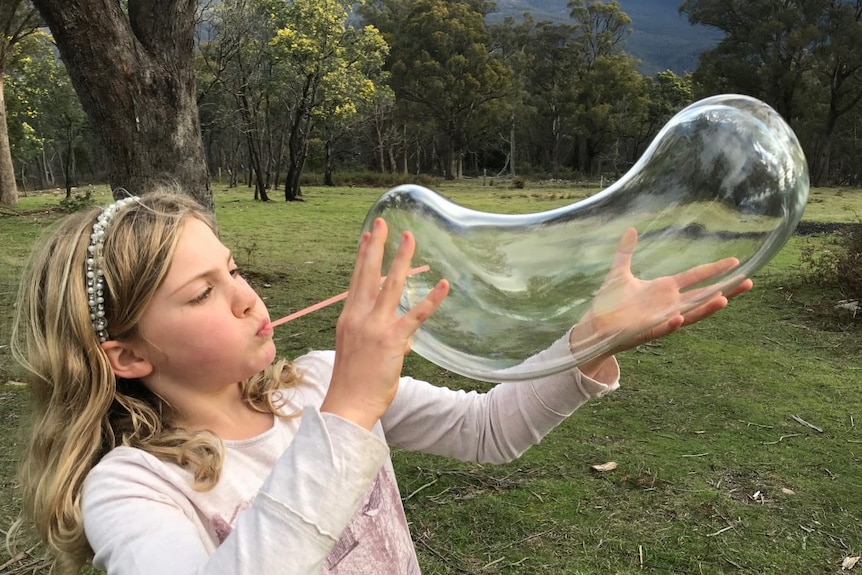 A girl blows air into a glass bubble through a straw against a bushy background.