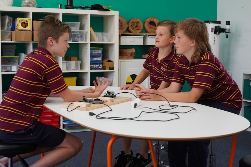 Three young boys wearing striped polo shirts and blue shorts sit around a table with a technical device and wires sitting on it