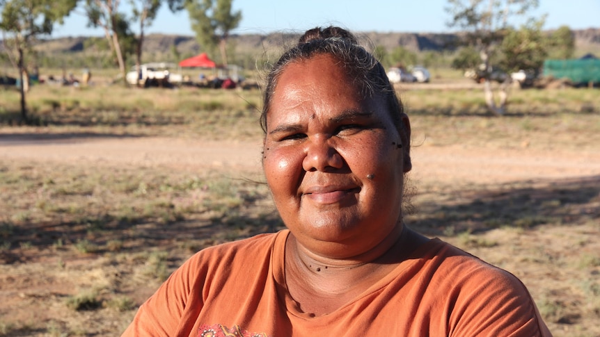 A women smiling at the camera with bush land and tents in the scenery behind her.