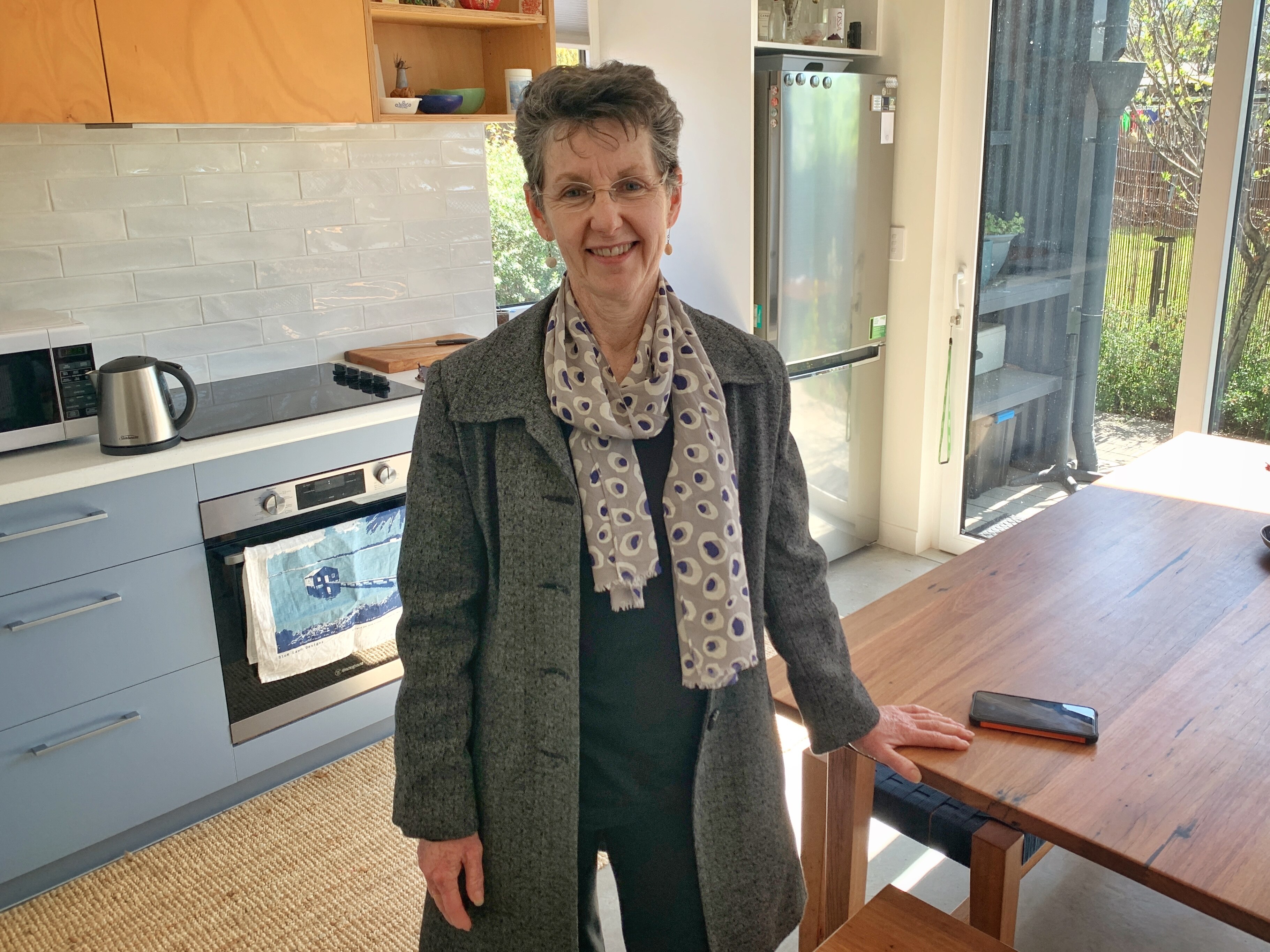 A woman standing in a kitchen in a grey blazer smiles. 