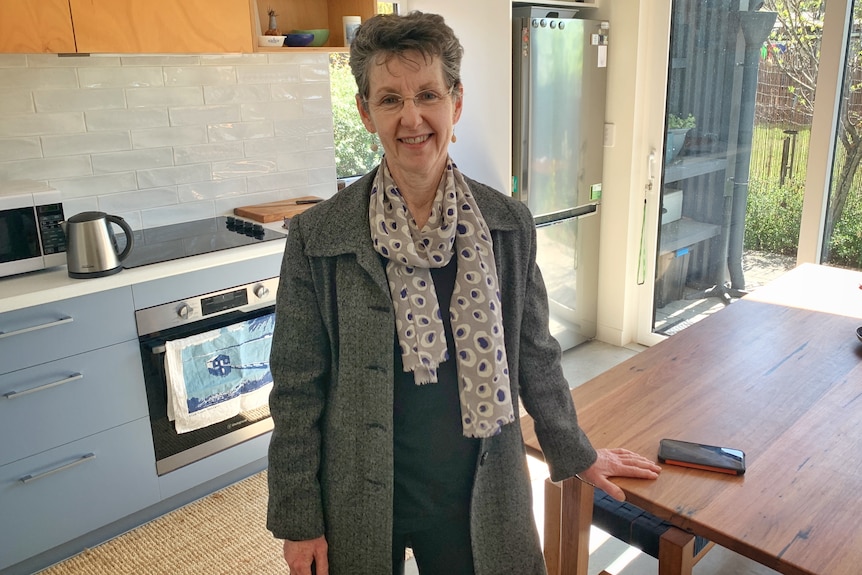 A woman standing in a kitchen in a grey blazer smiles. 