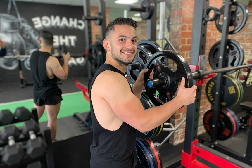 Andrew Kormas smiling while putting a 10kg weight on a bar near a squat rack in the gym.