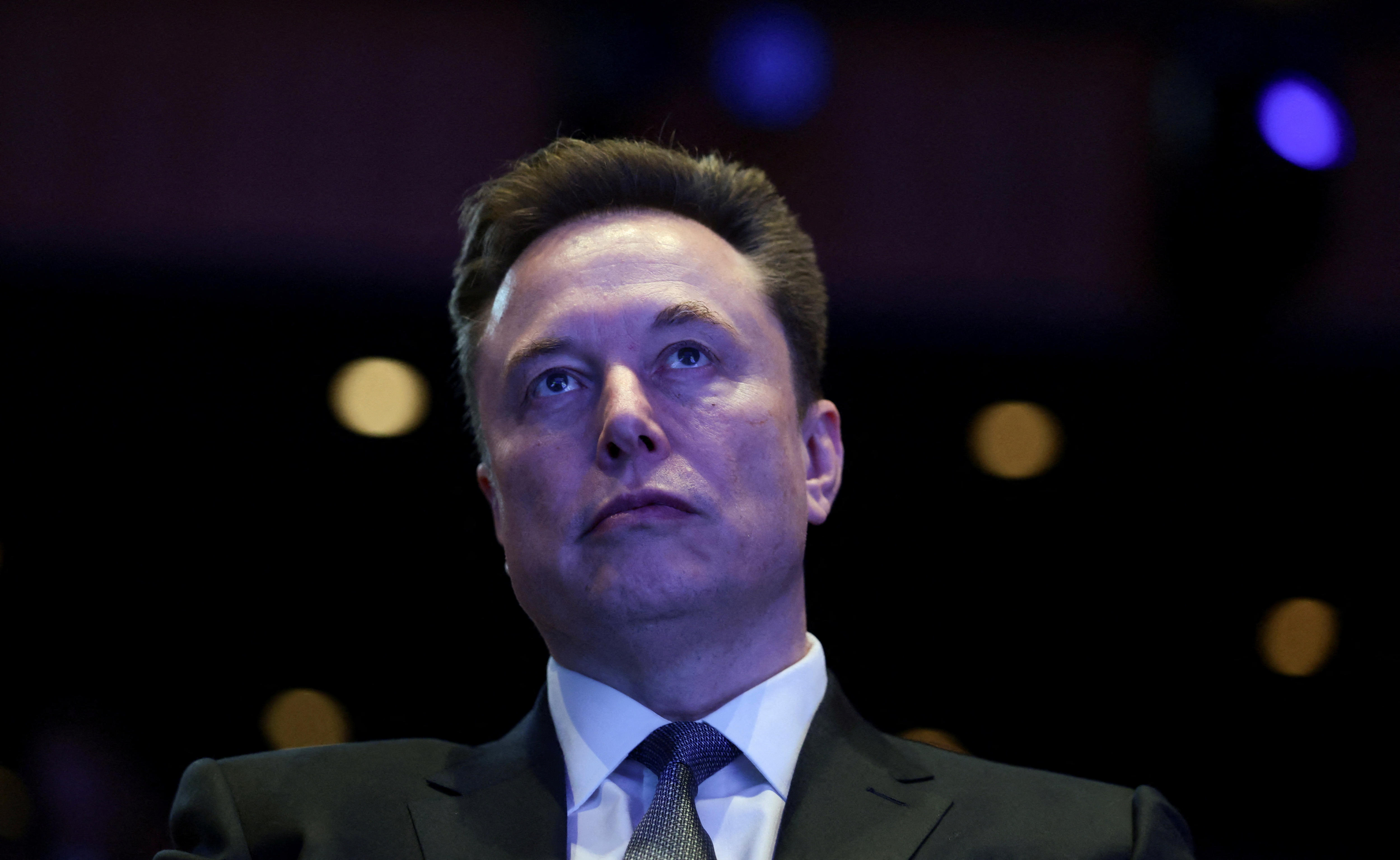 Elon Musk wearing a suit behind a dark backdrop.