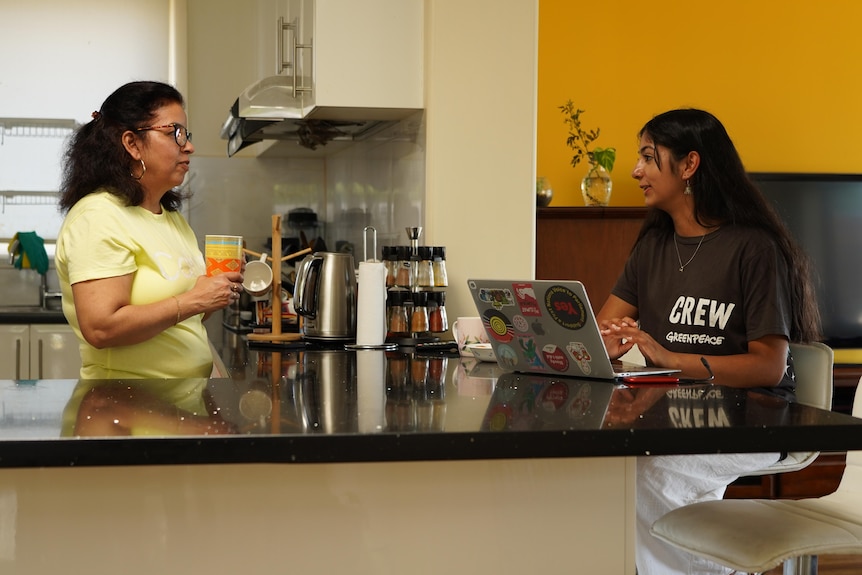 A teenager sits at a laptop at kitchen bench while her mum stands with a coffee cup across the bench from her