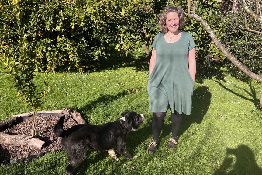 Leah is standing on grass in her garden in a green wool dress with a dog in front of her