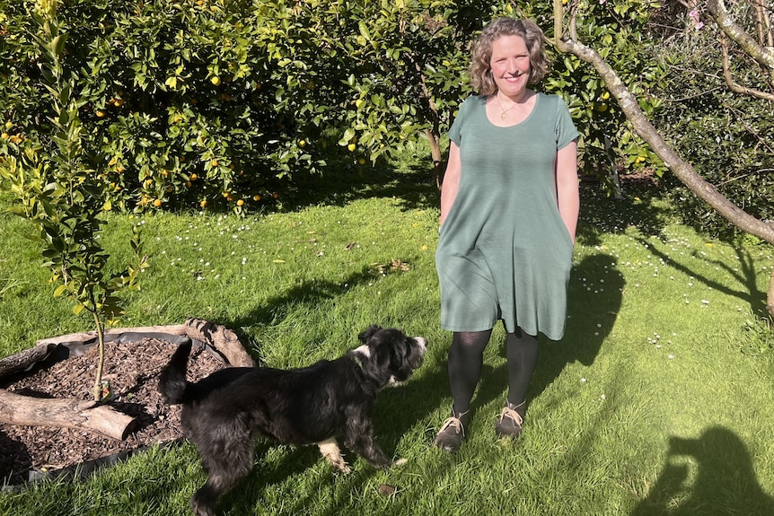 Leah is standing on grass in her garden in a green wool dress with a dog in front of her