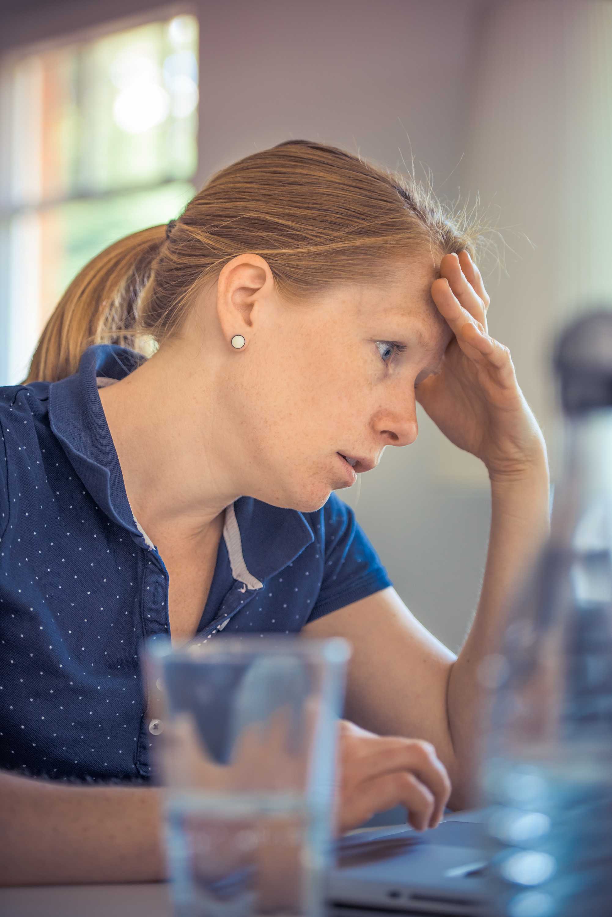 A woman in a blue shirt seated at a computer holding her head as a sign of stress.