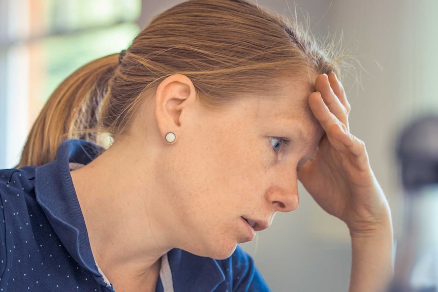 A woman in a blue shirt seated at a computer holding her head as a sign of stress.