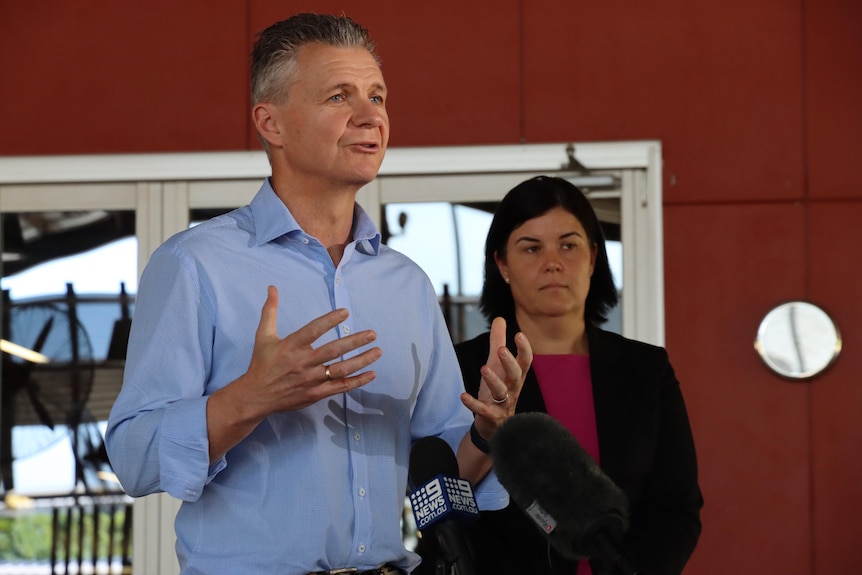 A man with grey hair and wearing a blue dress shirt is speaking and gesturing with his hands., Natasha Fyles stands behind him