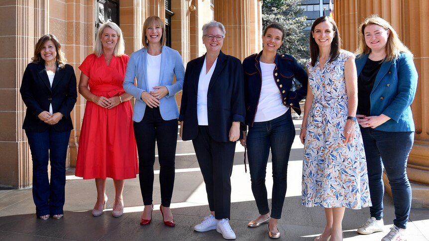 Seven women in a row under a sandstone portico. Five of the seven are wearing trousers, the other two dresses. All are smiling.