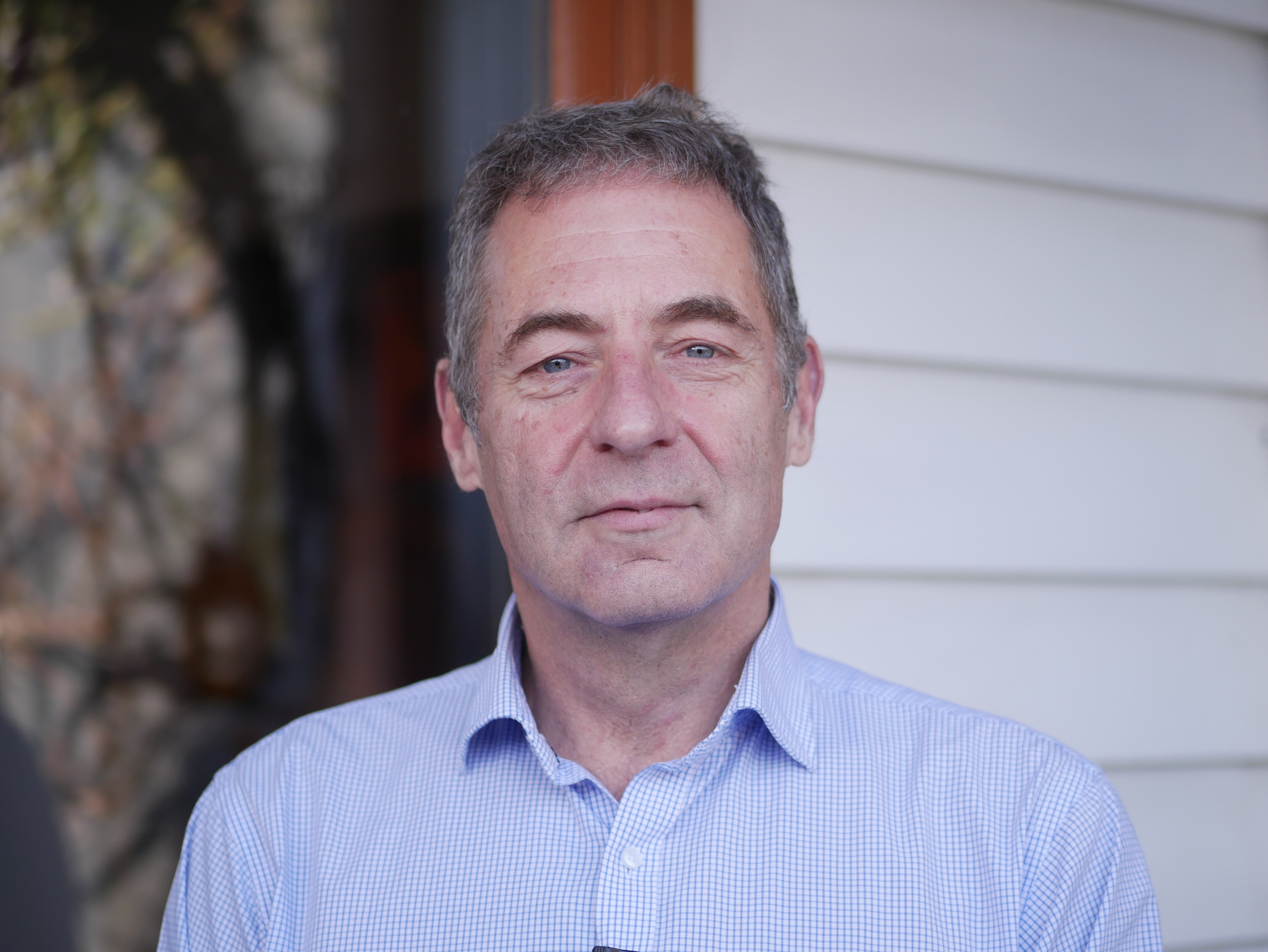 Portrait of a greying, blue-eyed man wearing a blue shirt outside a weatherboard house