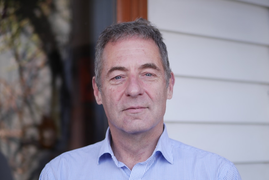 Portrait of a greying, blue-eyed man wearing a blue shirt outside a weatherboard house