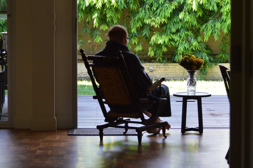 An elderly man in silhouette sitting in a chair looking at trees.