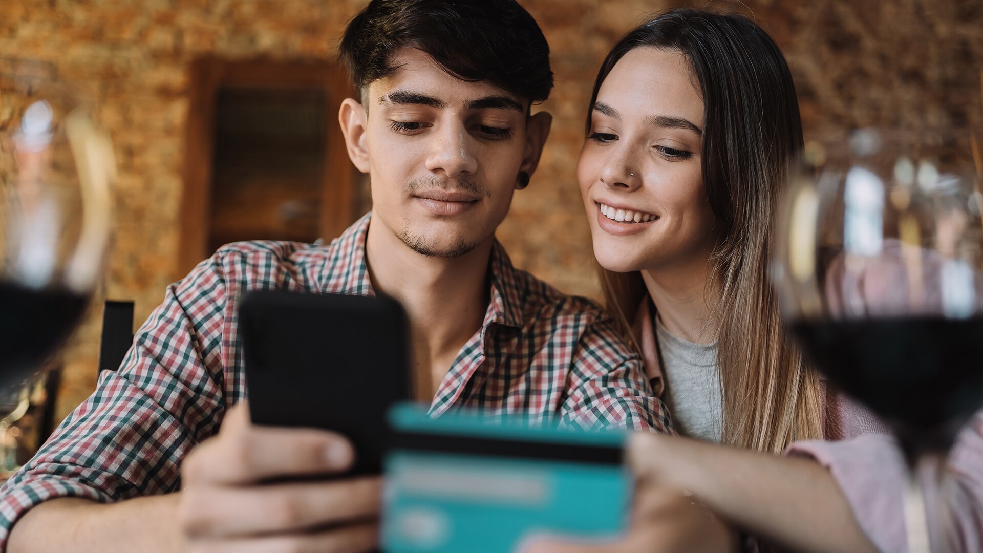 Happy couple sitting down looking at phone together. A blue bank card and two glasses of wine in the foreground out of the focus