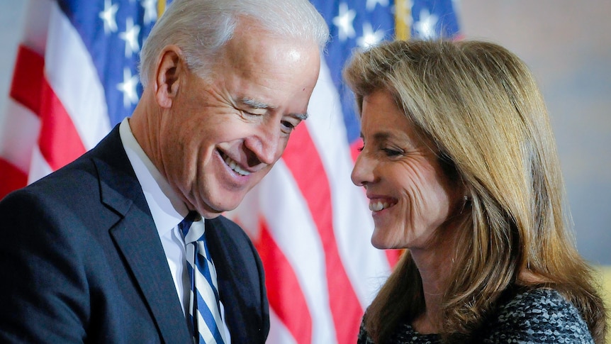 Joe Biden and Caroline Kennedy grinning at each other in front of a US flag. 