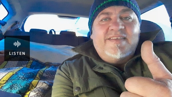 selfie of man in blue beanie with grey goatee beard raising one thumb in a car with bedding in teh back. Has Audio.