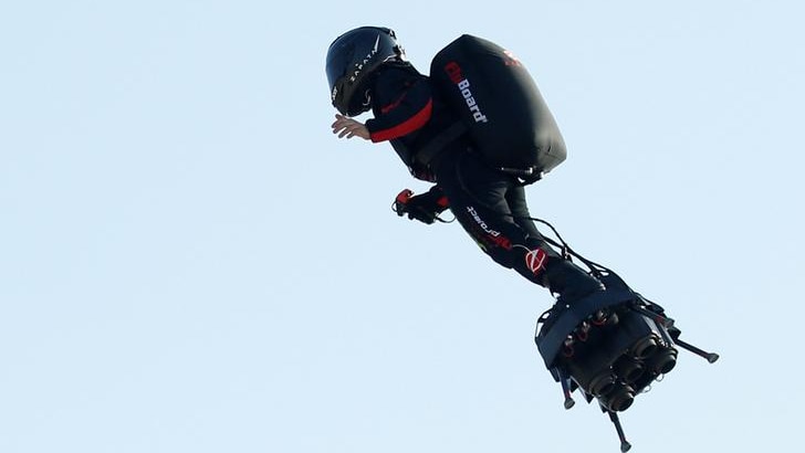 Franky Zapata wears a black suit with red embellishments as he soars in the sky on his Flyboard. The sky above is blue.