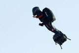 Franky Zapata wears a black suit with red embellishments as he soars in the sky on his Flyboard. The sky above is blue.