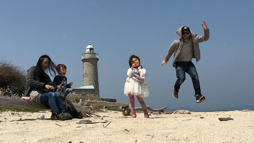 A family on the beach with the father jumping in the air, the daughter giving a peace sign, and the mother and son smiling