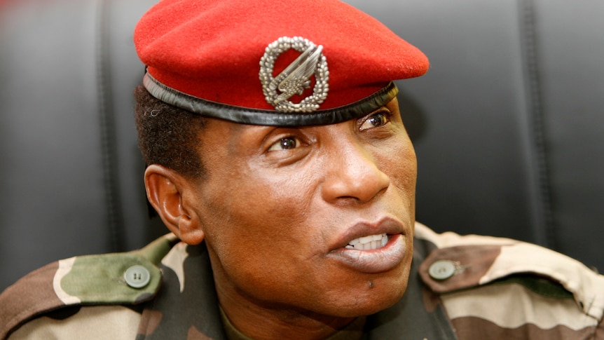 A middle-aged African man in a military uniform with a red beret speaks while seated in a leather chair.